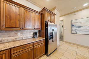 Kitchen with ornamental molding, brown cabinetry, light stone countertops, tasteful backsplash, and stainless steel refrigerator with ice dispenser