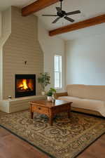 Living area with a large fireplace, ceiling fan, beamed ceiling, and wood finished floors