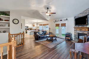 Living area featuring lofted ceiling, wood finished floors, a stone fireplace, and ceiling fan with notable chandelier