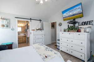 Bedroom with light wood-style flooring, a barn door, ensuite bathroom, lofted ceiling, and a ceiling fan