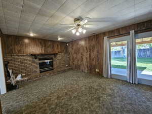 Unfurnished living room with carpet, wood walls, visible vents, a brick fireplace, and ceiling fan