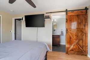 Bedroom featuring a barn door, ensuite bathroom, ceiling fan, light wood-style flooring, and baseboards