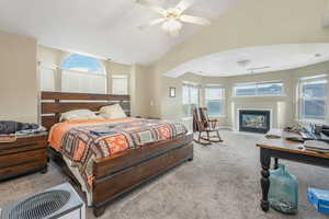 Carpeted bedroom featuring a fireplace with flush hearth, lofted ceiling, arched walkways, baseboards, and ceiling fan