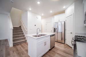 Kitchen featuring a sink, an island with sink, light wood-style floors, stainless steel appliances, and white cabinetry