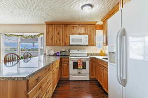 Kitchen with brown cabinetry, white appliances, stone countertops, a peninsula, and dark wood-style flooring