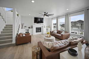 Living room with stairway, a ceiling fan, a glass covered fireplace, light wood-style flooring, and recessed lighting