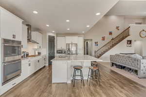 Kitchen with wall chimney exhaust hood, stainless steel appliances, light wood finished floors, an island with sink, and a breakfast bar area
