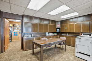 Kitchen featuring wooden walls, brown cabinetry, washer / dryer, a paneled ceiling, and light colored carpet