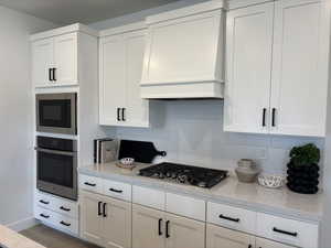 Kitchen with decorative backsplash, custom exhaust hood, white cabinetry, and appliances with stainless steel finishes