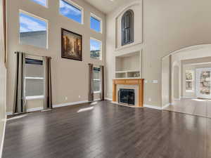 Unfurnished living room with built in shelves, arched walkways, a tiled fireplace, a high ceiling, and dark wood-type flooring