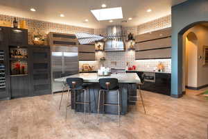 Kitchen featuring wall chimney exhaust hood, arched walkways, a skylight, stainless steel built in fridge, and a breakfast bar area