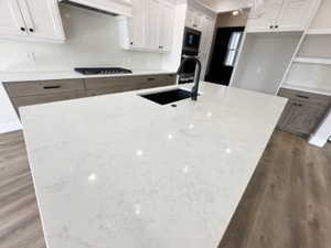 Kitchen featuring a sink, black stovetop, ventilation hood, wood finished floors, and a kitchen island with sink