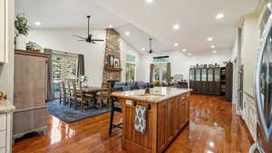 Kitchen with ceiling fan, open floor plan, a stone fireplace, brown cabinetry, and high vaulted ceiling