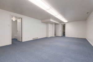 Finished below grade area with a textured ceiling, dark colored carpet, visible vents, and baseboards