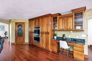 Kitchen featuring double oven, built in desk, baseboards, dark wood finished floors, and brown cabinetry