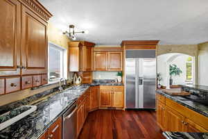 Kitchen with dark stone countertops, brown cabinetry, stainless steel appliances, a sink, and dark wood-type flooring