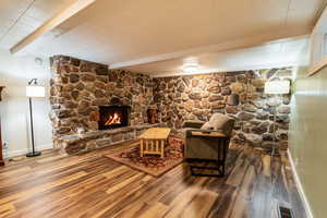 Living room featuring baseboards, wood finished floors, a stone fireplace, beam ceiling, and visible vents
