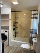 Bathroom featuring a textured ceiling, washer / clothes dryer, shower / bath combo with shower curtain, toilet, and wood finished floors