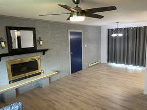 Unfurnished living room with a brick fireplace, baseboard heating, wood finished floors, and ceiling fan with notable chandelier