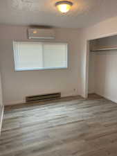 Unfurnished bedroom featuring a textured ceiling, a baseboard heating unit, a closet, wood finished floors, and a wall mounted air conditioner