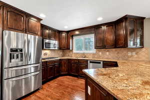 Kitchen featuring dark brown cabinets, wood finished floors, light stone countertops, decorative backsplash, and appliances with stainless steel finishes