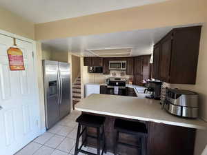 Kitchen with stainless steel appliances, light tile patterned flooring, dark brown cabinetry, a peninsula, and light countertops