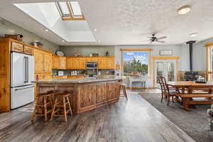 Kitchen with an island with sink, dark wood-style floors, a wood stove, appliances with stainless steel finishes, and ceiling fan