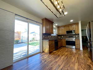 Kitchen featuring dark wood finished floors, dark countertops, decorative backsplash, brown cabinetry, and appliances with stainless steel finishes