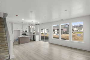 Living room with recessed lighting, light wood finished floors, and large windows leading into the dining room and kitchen with a sink, an inviting chandelier, and island
