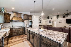 Kitchen featuring custom range hood, appliances with stainless steel finishes, ceiling fan, light stone countertops, and vaulted ceiling