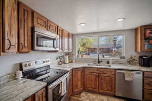 Kitchen with light stone counters, stainless steel appliances, brown cabinets, a sink, and a textured ceiling