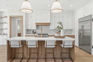 Kitchen with white cabinets, light wood-type flooring, decorative backsplash, a kitchen island with sink, and stainless steel built in refrigerator