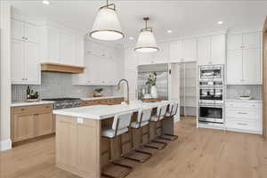 Kitchen featuring decorative light fixtures, light wood-type flooring, decorative backsplash, a kitchen island with sink, and appliances with stainless steel finishes