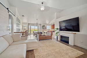 Living area with a barn door, a glass covered fireplace, vaulted ceiling, wood finished floors, and ceiling fan with notable chandelier