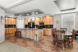 Kitchen featuring a kitchen island with sink, tasteful backsplash, wood finished floors, black appliances, and a raised ceiling