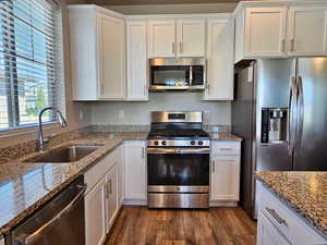Kitchen featuring dark wood-style flooring, appliances with stainless steel finishes, light stone countertops, a sink, and white cabinets