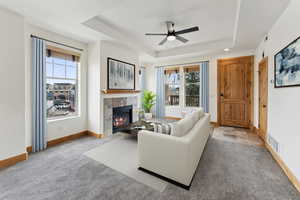 Carpeted living area featuring a tiled fireplace, a raised ceiling, ceiling fan, and a healthy amount of sunlight