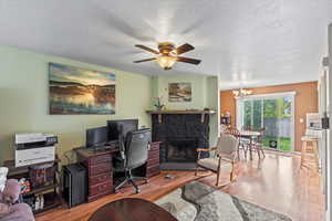 Office space featuring a textured ceiling, a stone fireplace, wood finished floors, and ceiling fan with notable chandelier