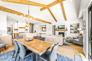 Dining room with a warm lit fireplace, an inviting chandelier, light wood-style flooring, recessed lighting, and vaulted ceiling with beams