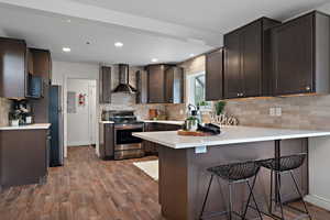 Kitchen featuring wood finished floors, decorative backsplash, stainless steel appliances, wall chimney range hood, and a peninsula