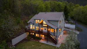 Rear view of house featuring a wooded view, concrete driveway, a deck, a patio area, and a shingled roof