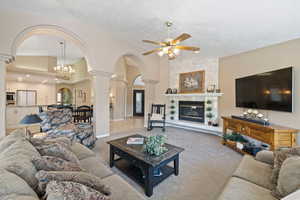 Carpeted living room featuring decorative columns, a textured ceiling, a high end fireplace, arched walkways, and ceiling fan with notable chandelier