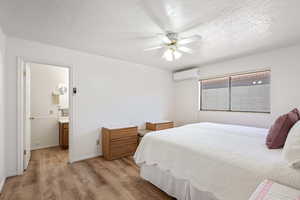 Bedroom featuring a ceiling fan, a textured ceiling, light wood-type flooring, an AC wall unit, and ensuite bathroom