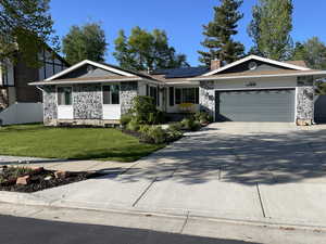 Single story home with stone siding, a front yard, roof mounted solar panels, an attached garage, and driveway