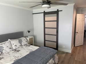 Bedroom featuring a ceiling fan, a barn door, wood finished floors, baseboards, and ornamental molding