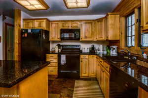 Kitchen with brown cabinetry, a sink, dark stone countertops, and black appliances
