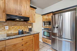 Kitchen with light stone counters, tasteful backsplash, stainless steel appliances, and under cabinet range hood