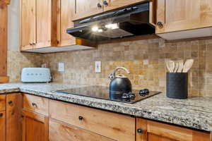 Kitchen featuring decorative backsplash, under cabinet range hood, black electric stovetop, and light stone countertops