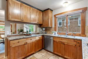 Kitchen featuring brown cabinetry, backsplash, dishwasher, a sink, and light stone counters