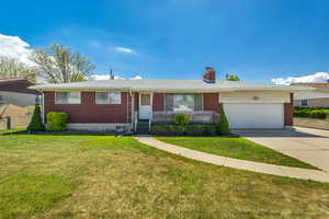 Single story home featuring an attached garage, a front lawn, brick siding, a chimney, and driveway